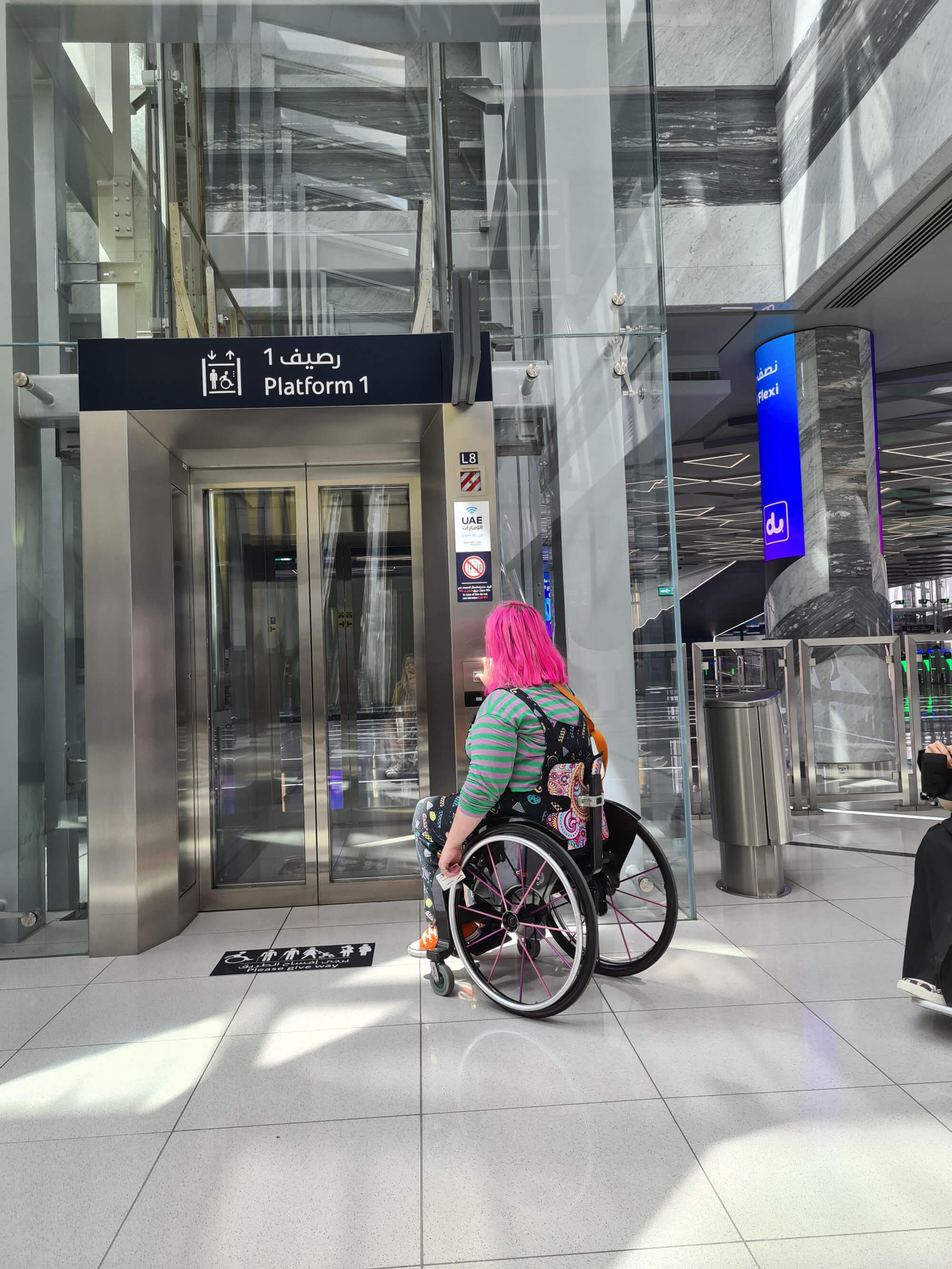 Emily Yates of Mima in her wheelchair in front of the doors to a large steel and glass elevator, with a sign saying 
