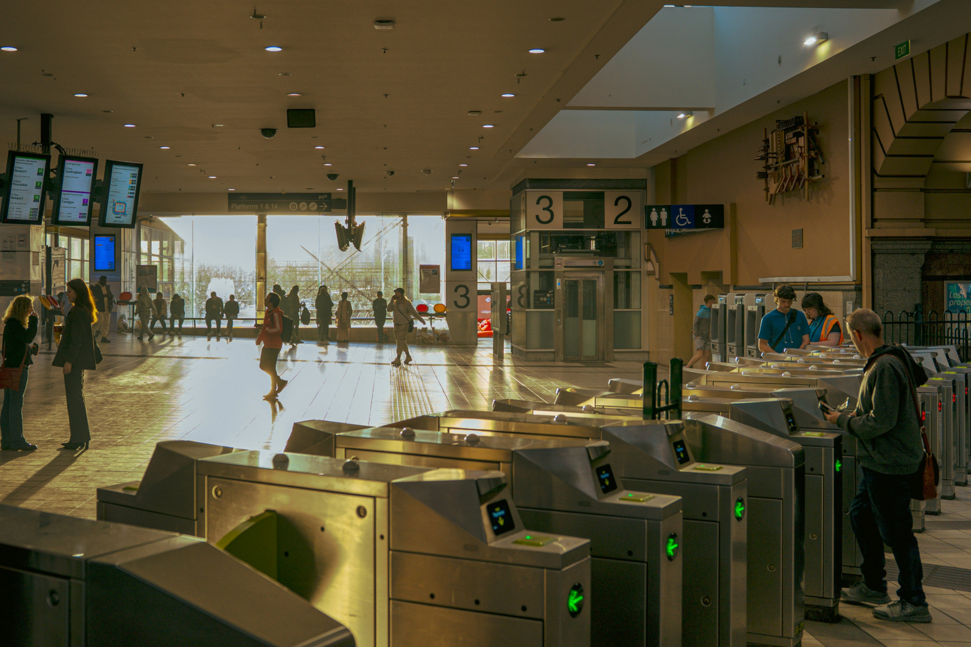 Large, sunlit train station concourse with ticket gates in the foreground and people walking and waiting near platforms in the background.