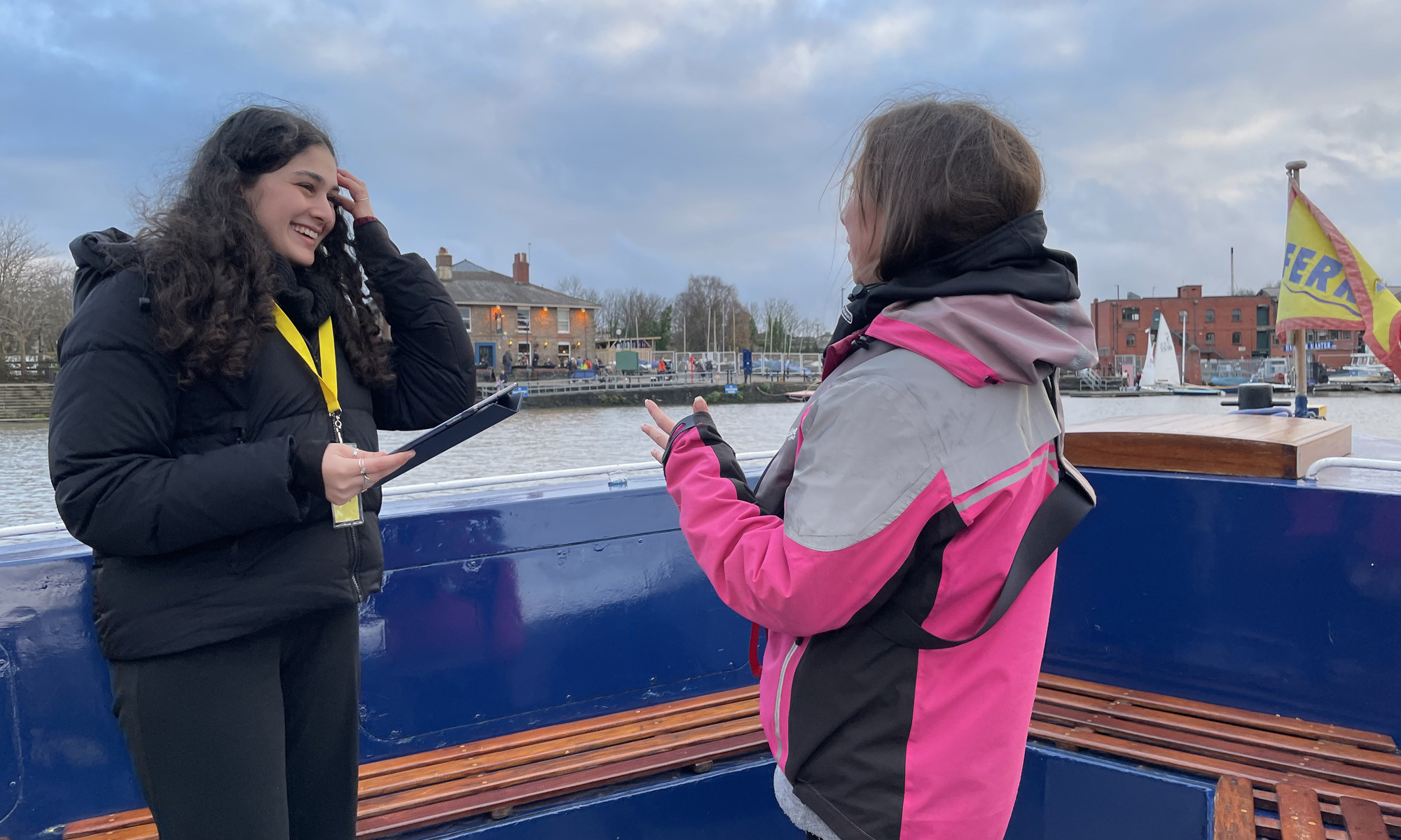 Two women talking on a ferry boat in Bristol. One, a Mima team member holds a clipboard and wears a yellow lanyard, interviewing the other who wears a pink and grey jacket. In the background, buildings, trees, and boats are visible across a river under a cloudy sky.