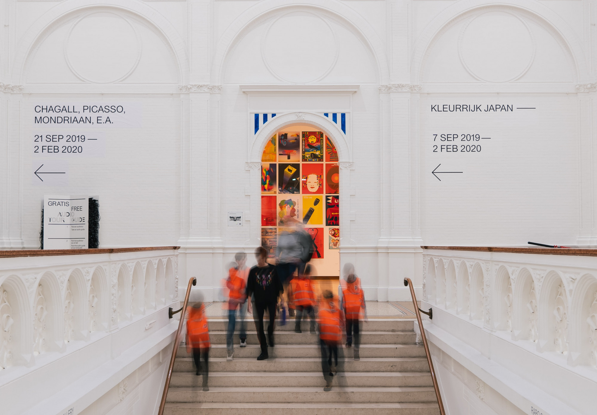 Blurred outlines of school children in orange high vis jackets and an adult walk down a central staircase in a museum, with a white wall and doorway behind them with exhibition signage directions on the wall.