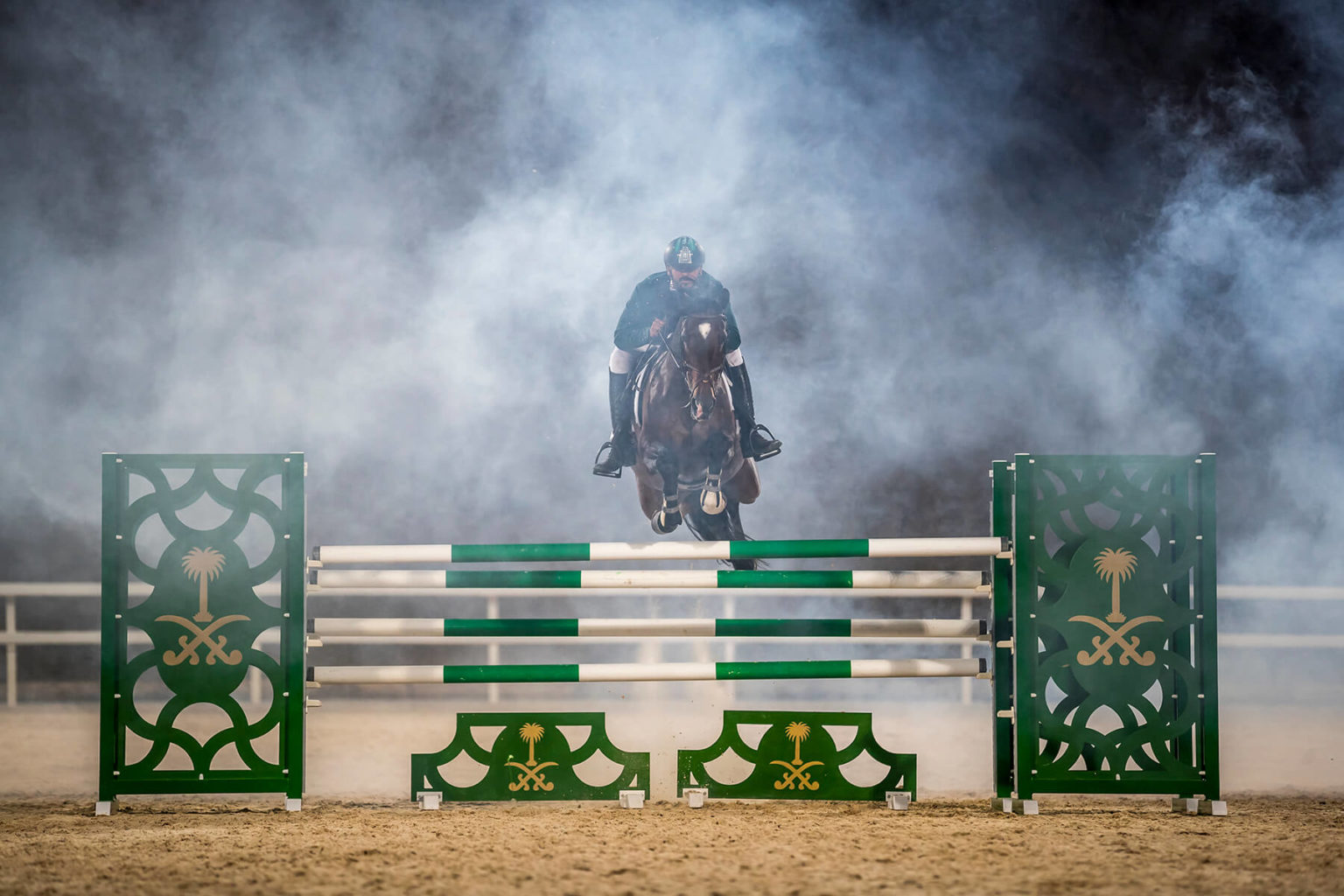 A white jockey with a moustache is showjumping on a brown horse. The two are in mid air over a white, green and gold hurdle. Smoke is behind them, and a sandy floor is beneath them.