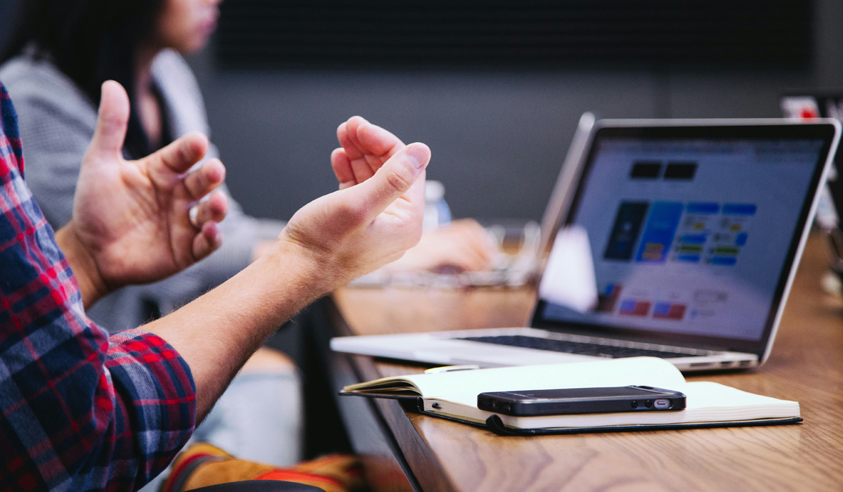 Close-up of a person gesturing with their hands while sitting at a conference table with a laptop displaying a colorful dashboard or website, a notebook, and a smartphone. Another person is blurred in the background, suggesting a meeting or collaborative work setting.