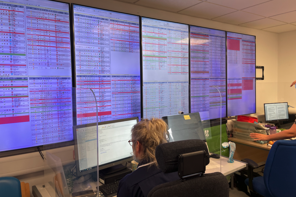 A close up of a control room that manages urgent referrals. Five large, vertical screens fill the image, and tables fill the screens with information colour coded in white, red and green. A staff member with glasses and blonde hair sits at their desk and faces the screens.