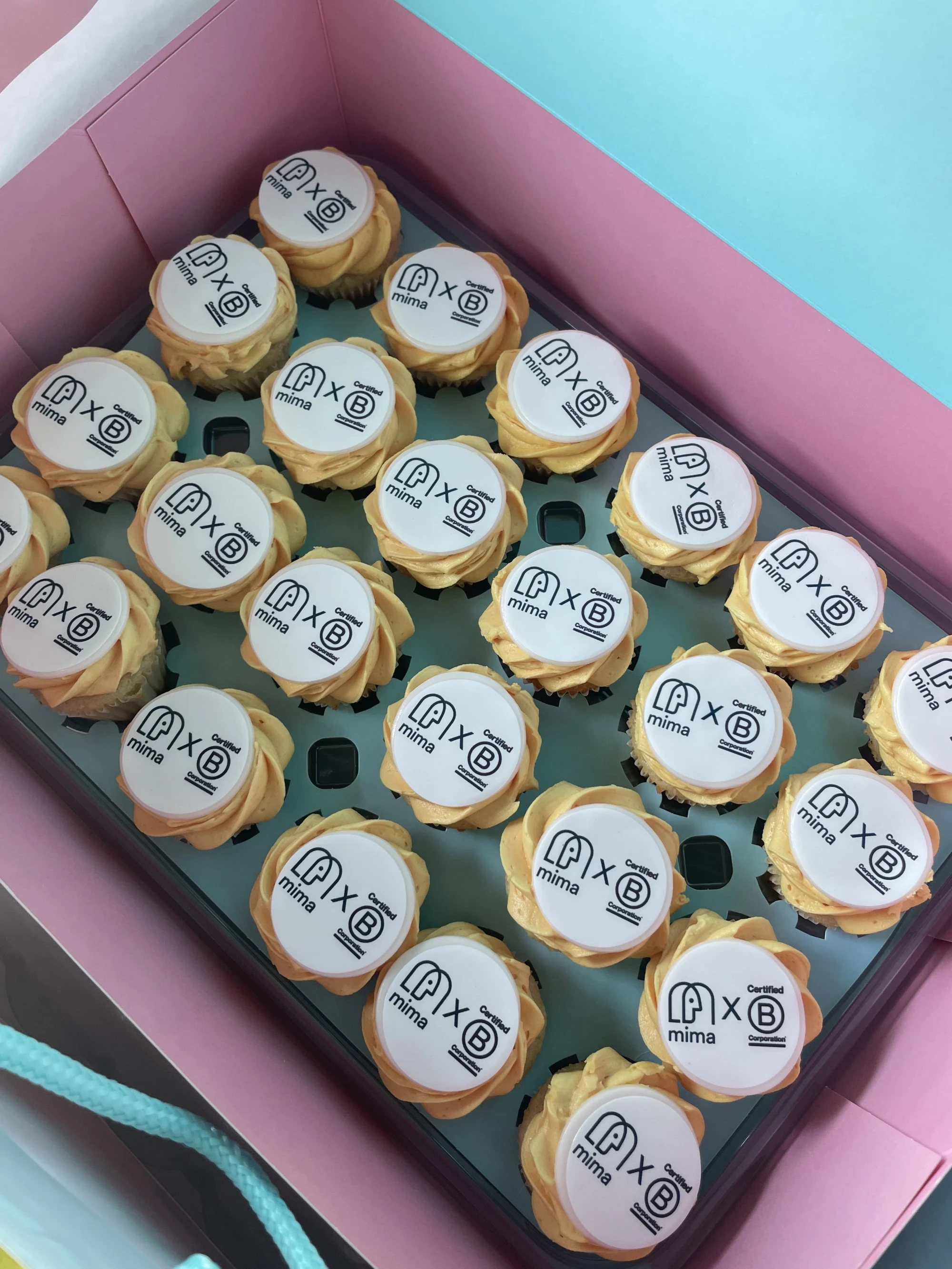 A photo of the inside of a box of cupcakes decorated with both the logos of Mima and B Corp