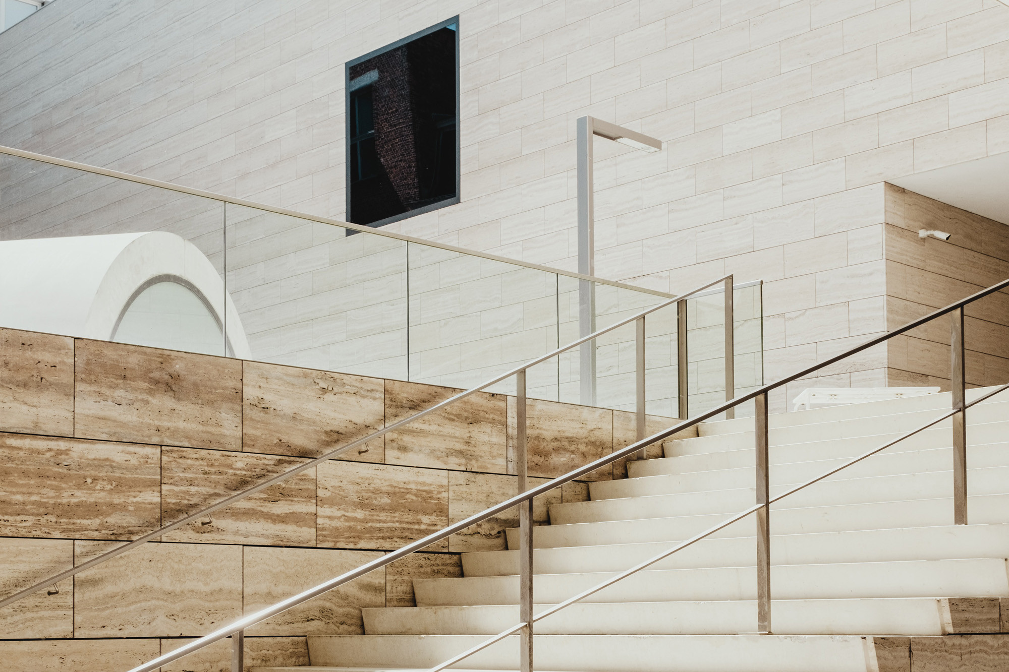 A minimalist white staircase alongside a stone wall, looking to an upper level in an open plan space, with a digital screen but no obvious wayfinding signage.