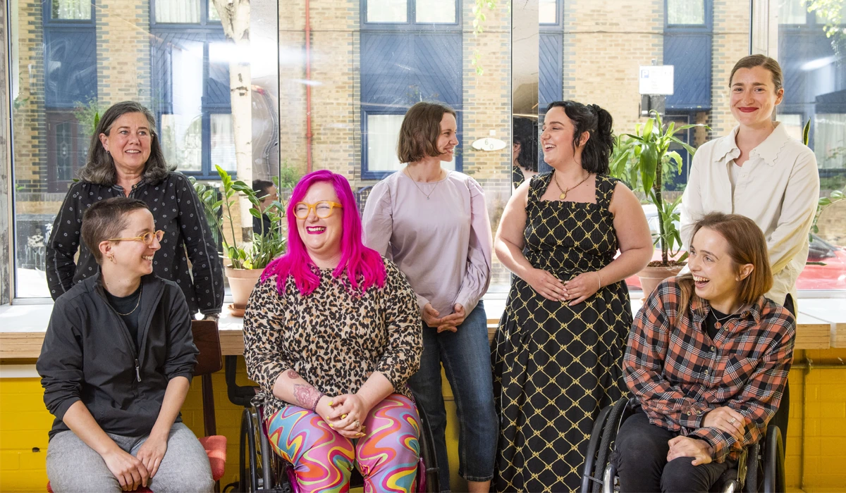 Mima's Accessibility & Inclusive Design Consultant team in a group shot together in a London office. They are smiling and laughing together.