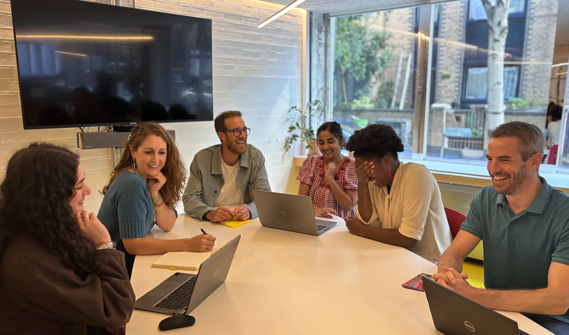 Six people sit around a table talking and laughing. A blank TV screen is on the wall behind them and they have laptops and notepads out on the table.