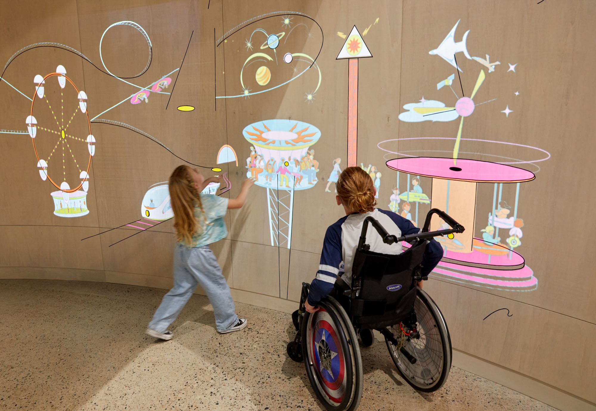 Two children, one a wheelchair user look up at colourful lit up projections onto a curved wooden wall within a gallery. The lit up drawings on the wall depict fairground rides including a carousel.