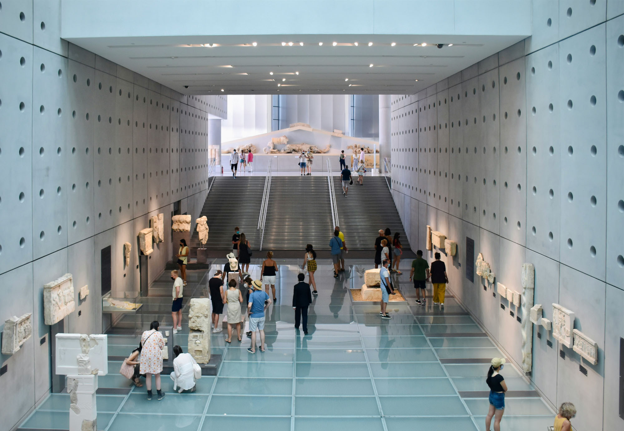 Visitors stand in groups looking at artefacts in a vast open museum. The floor is glass panelled, with concrete walls, displaying carved sculptures. In the view beyond is a wide staircase leading up to another level of the museum.