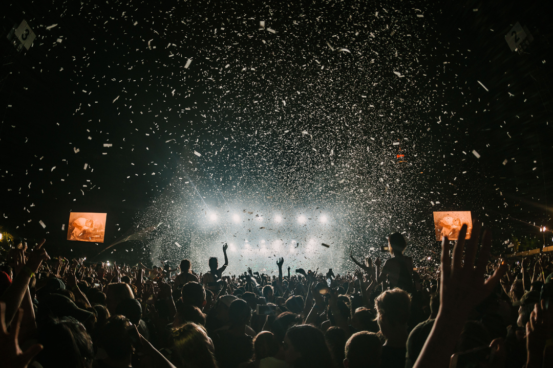 A large crowd of people is gathered at an outdoor concert at night. The stage is brightly lit with white lights beaming, and confetti is falling from above. Many attendees have their hands raised, and the scene captures the excitement of the event.