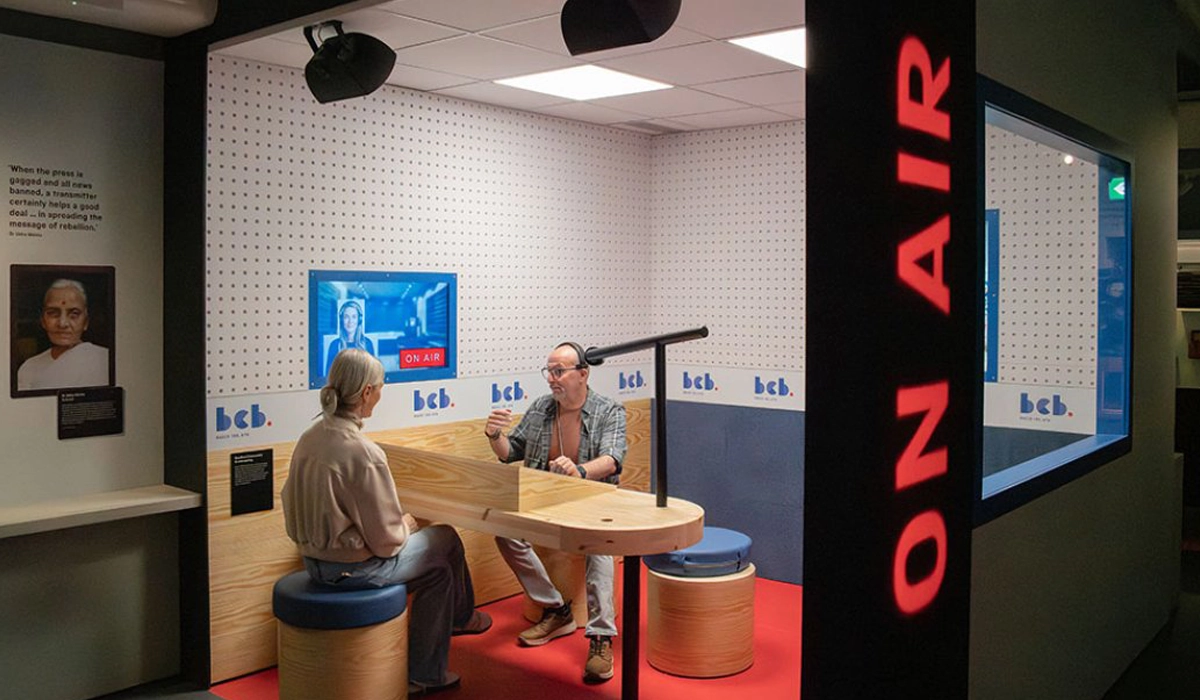 Two people sit inside a replica of a recording studio booth with the words 'on air' down the outside wall in red lettering, at the Sound + Vision galleries at the National Science museum.