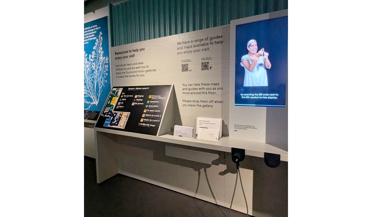 The Access Hub display at the Sound and Vision galleries showing resources to help visitors enjoy their visit. The display shows a tactile floorplan of the galleries alongside maps and guides that can be taken around the gallery. A digital screen shows a person using BSL to explain how to connect to BSL interpretation using QR codes around the exhibition.