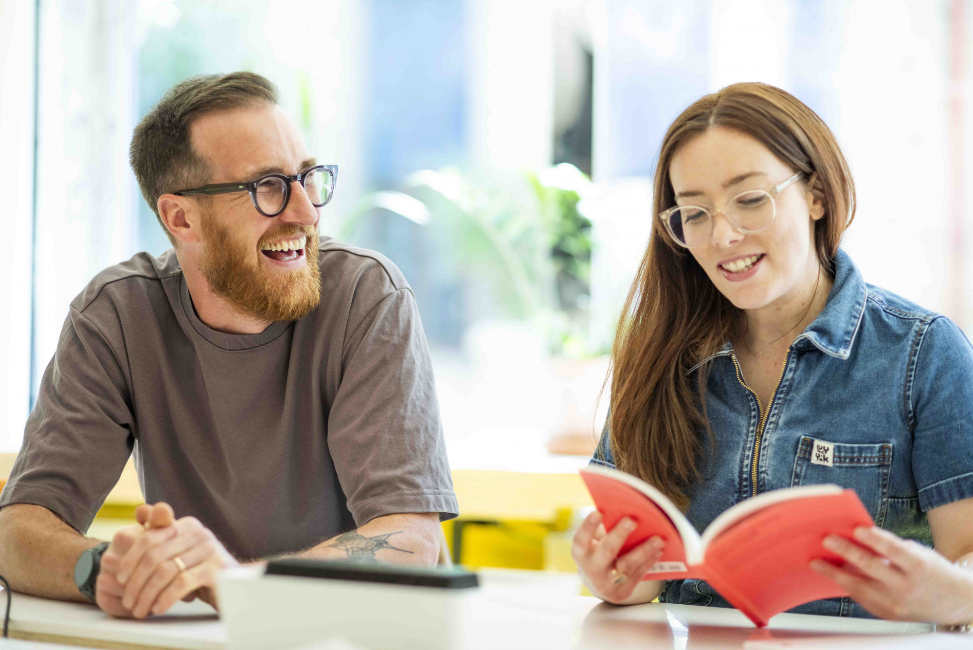 Two Mima team members sit at a table in a meeting room, Adam a white male with black glasses  is laughing and Alice, a white female with clear rimmed glasses, is reading a book with a red cover.