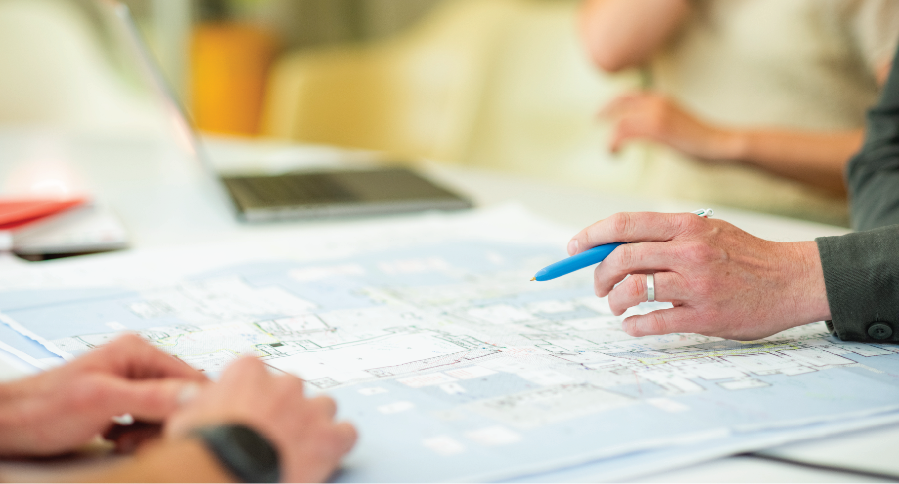 A hand holding a pen hovers over a map laid out across a table. People are sat around the table but only their hands are in view.