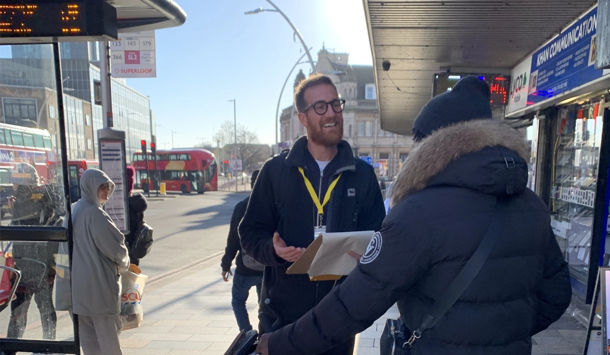 Adam, a member of the team at Mima holds a clipboard whilst interviewing a passenger at a London bus stop in a built up area.