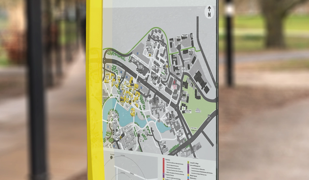 A close-up view of an outdoor wayfinding map display in a park or pedestrian area, showing a detailed layout of buildings, paths, green spaces, and water features. The map includes a “You Are Here” marker, a north arrow for orientation, and a color-coded legend at the bottom identifying different types of buildings or departments. The display is encased in a yellow frame, with blurred trees and pathways visible in the background.