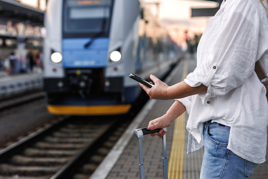 A person holding their phone and a suitcase is waiting on a station platform as a train approaches