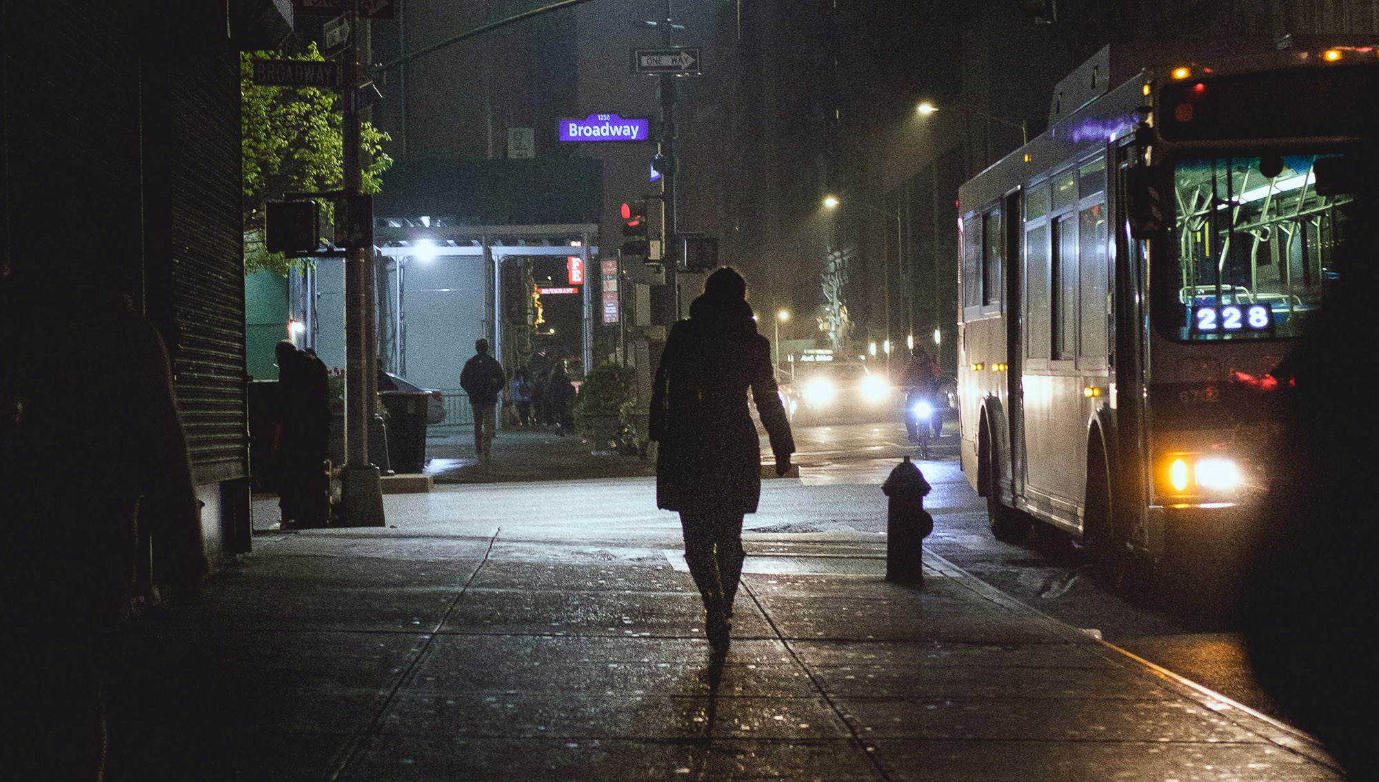 A figure walks on a pavement at night through a town area beside a road where a bus and motorcycle are passing. The back of the figure is in sight, with signage and street lights beyond.
