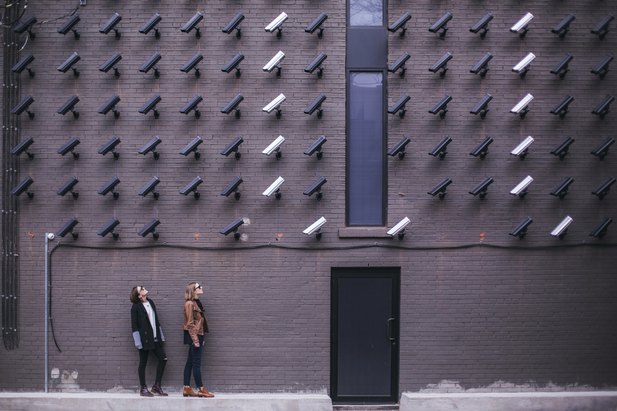 Two people stand looking up at a brown brick wall with lines of security cameras facing down towards them.