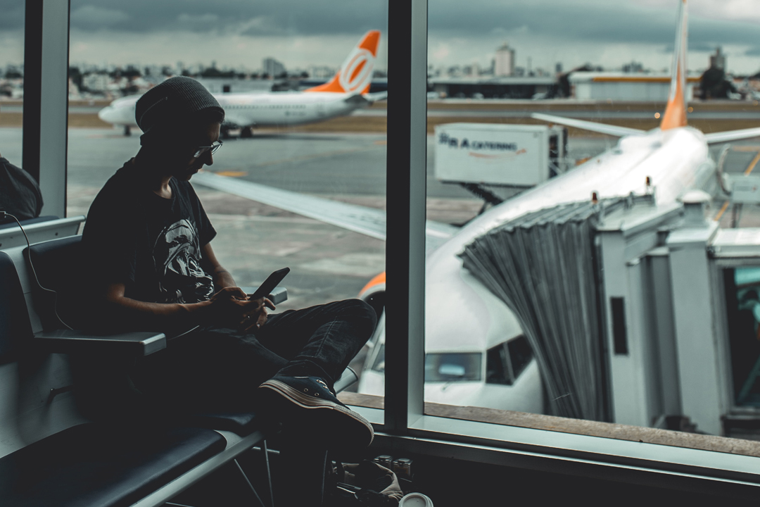 Person sitting in airport departure lounge beside a window. Through the window planes can be seen on the runway.