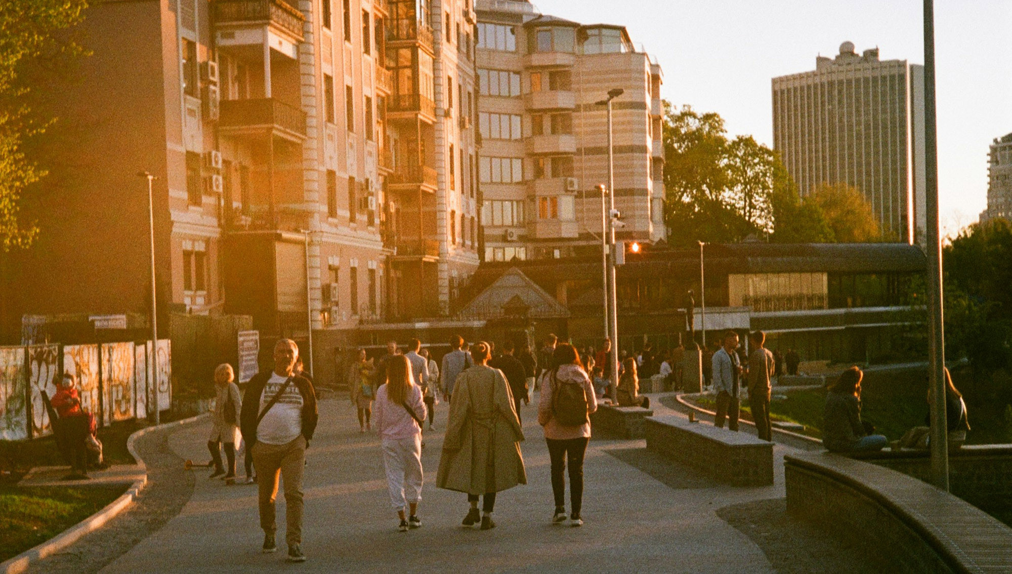 People walk along a curved path with bench seating to the side and grass areas. Sunlight shines onto the front of buildings near by.