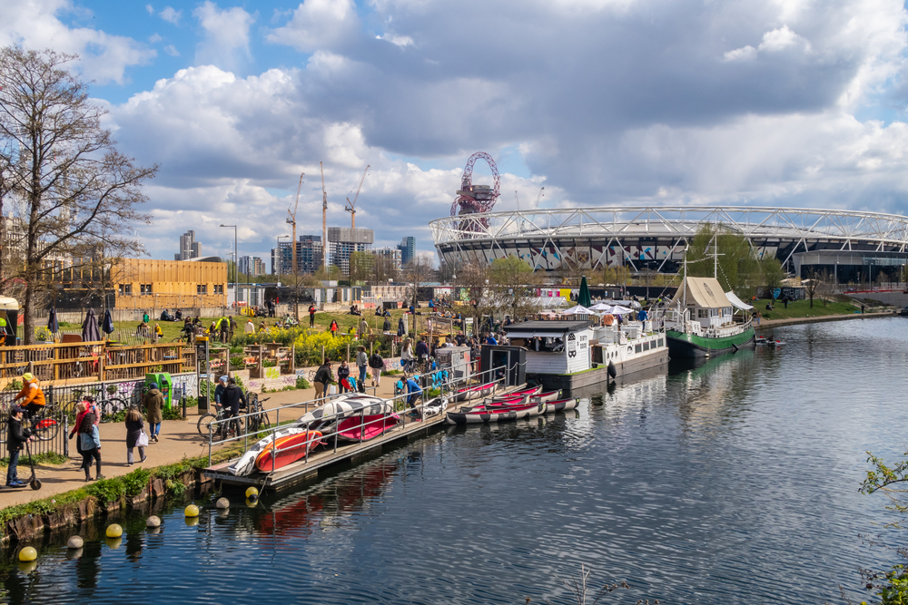People outside canal bars in Hackney Wick in London on a sunny day. In the background you can see the West Ham Stadium.