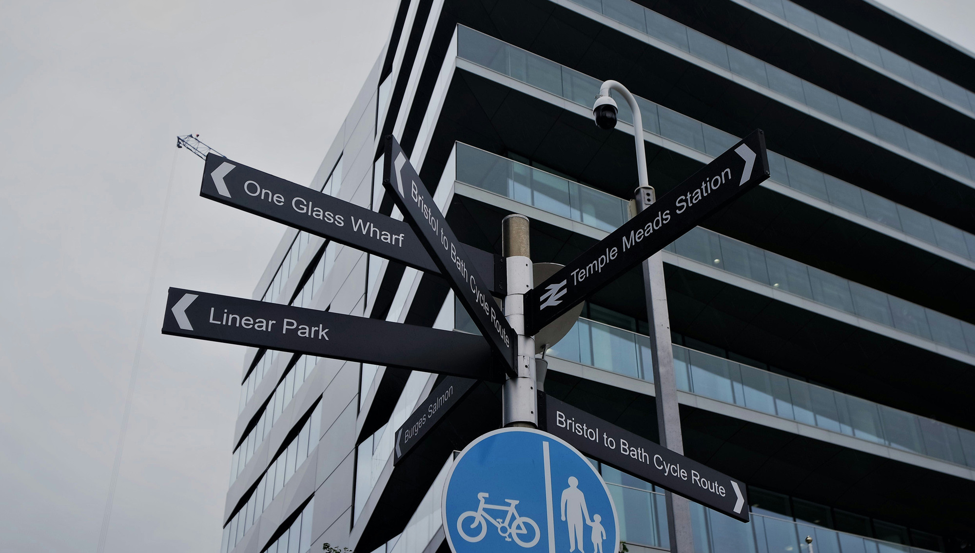 A directional signpost in Bristol with white writing on black rectangular directional signs. Signs direct to Temple Meads Station, Linear Park and Bristol to Bath cycle route. The sign also includes a blue circle with a symbol of a bicycle and a pedestrian.