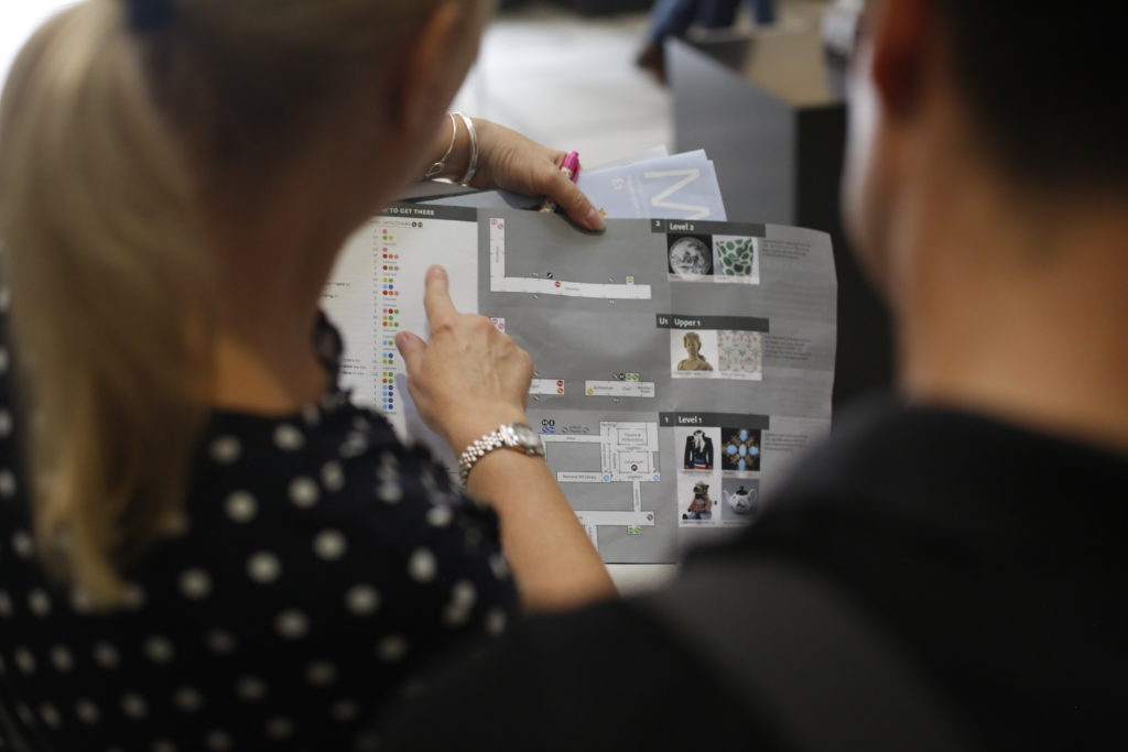 Two white people frame the image, with the close up of a grey and white map in the centre. The woman, who has blonde hair and a polka dot top, points at an element on the map.