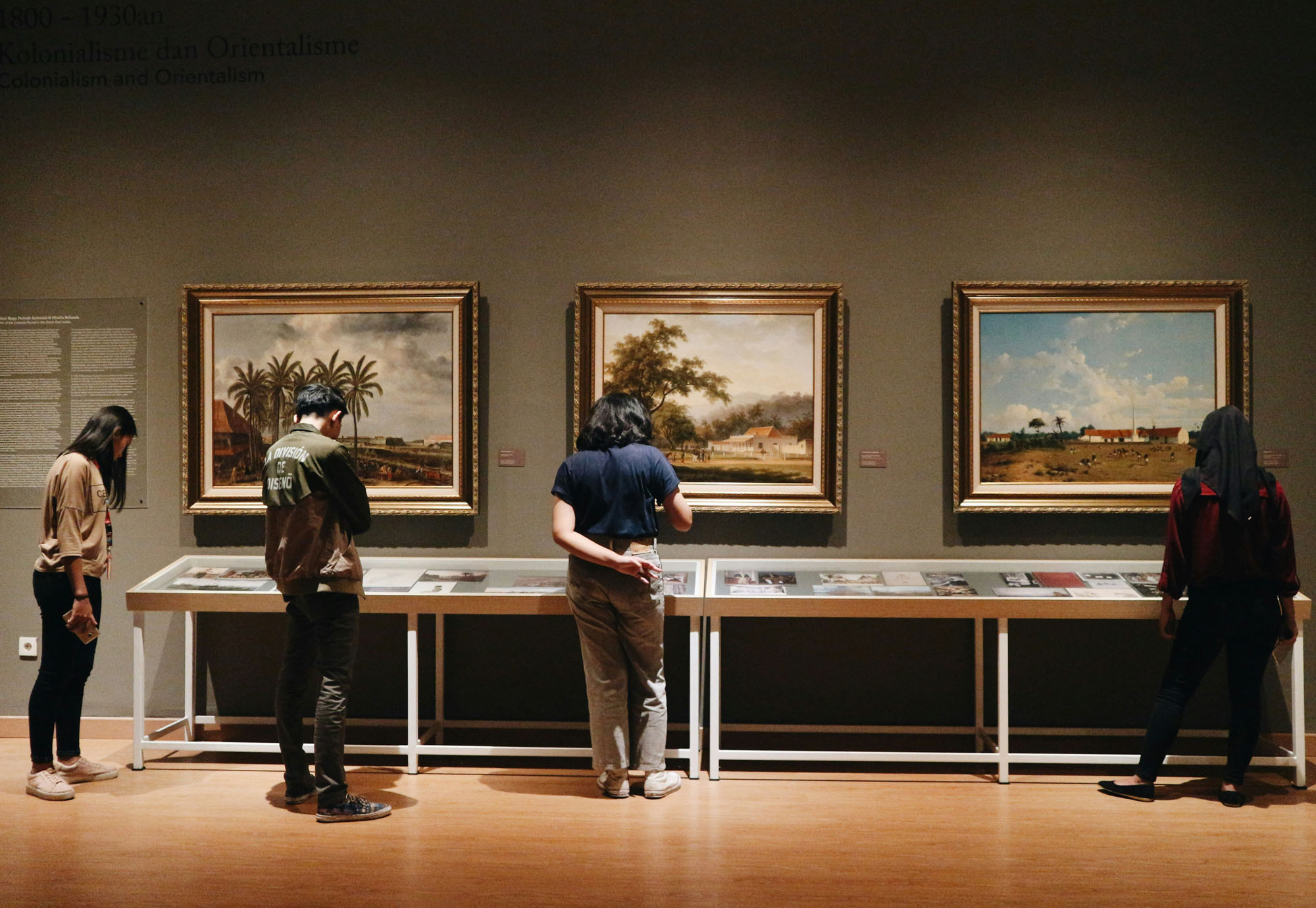 Four people stand with their backs turned away, looking down at an exhibit table within a museum. On the wall are three paintings of landscapes.