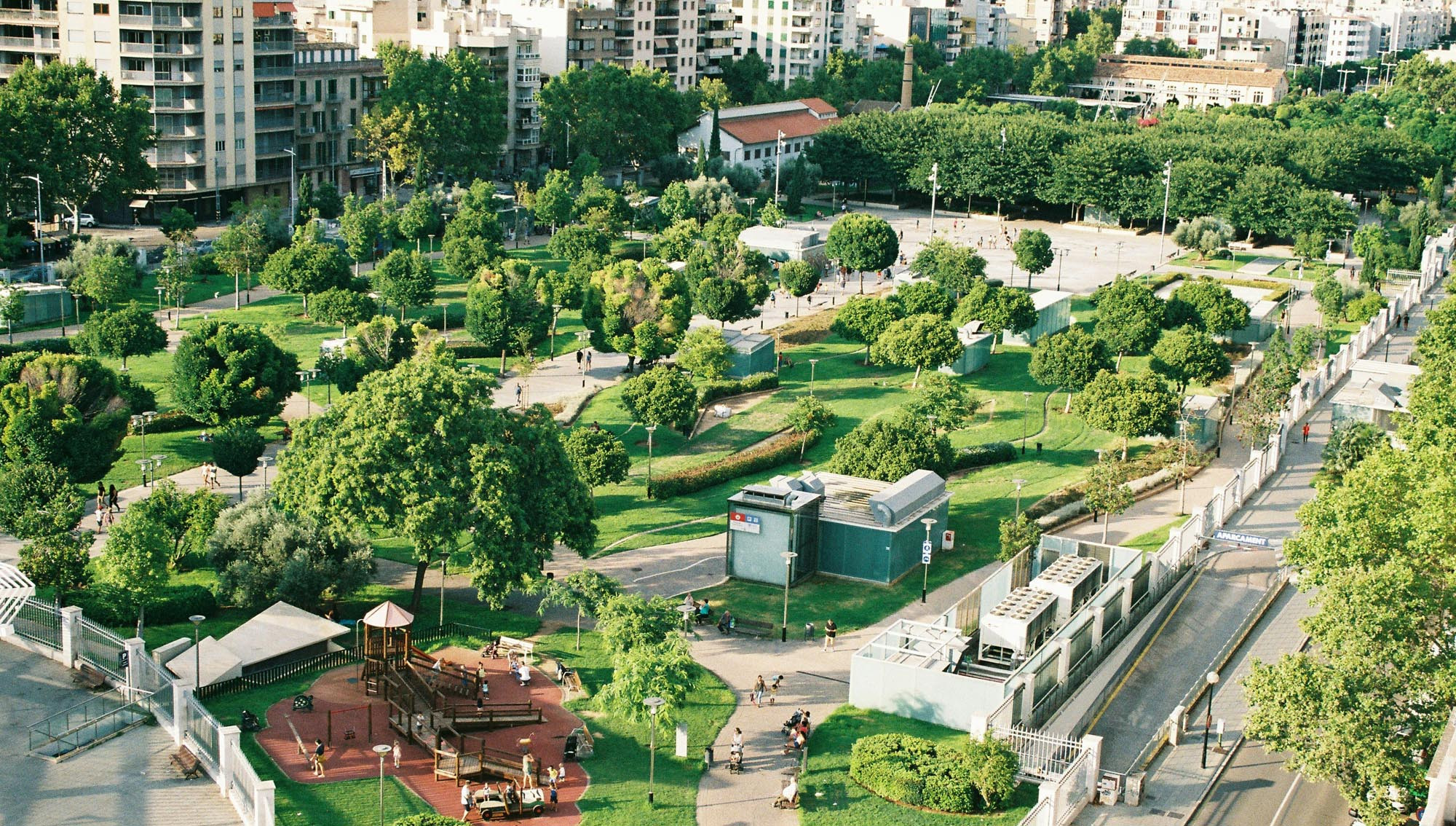 A green fenced off park area in the middle of a city with surrounding city skyscrapers and builders. Trees are surrounding play areas with pathways weaving in between the green areas.