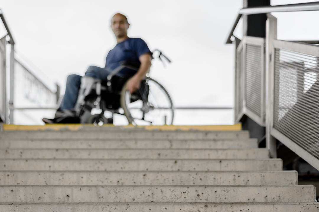 A man in a wheelchair is positioned at the top of a flight of stairs, with no visible ramp or accessible route in sight.