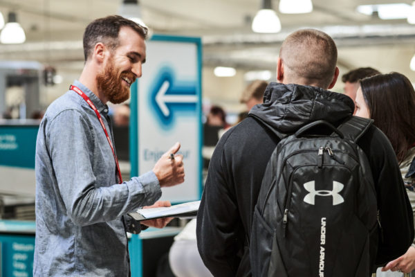 Adam, a Mima team member, wears a blue shirt and has a red beard. He is smiling whilst communicating with a passenger in John Lennon Airport. The passenger has his back to us, and wears black with a black rucksack on his back.