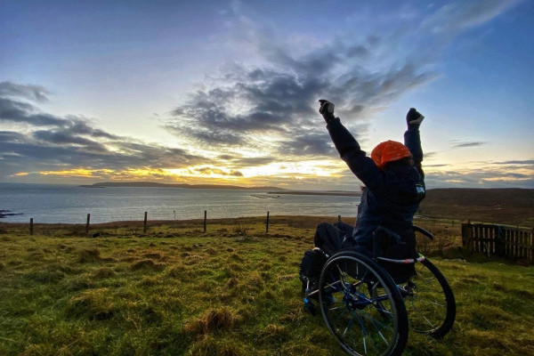 Wheelchair user, arms outstretched, watching the sunrise. Looking out towards Fetlar, Shetland, Scotland