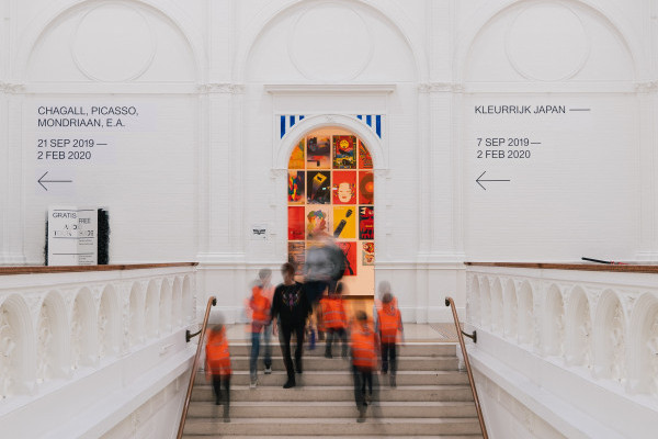Blurred outlines of school children in orange high vis jackets and an adult walk down a central staircase in a museum, with a white wall and doorway behind them with exhibition signage directions on the wall.
