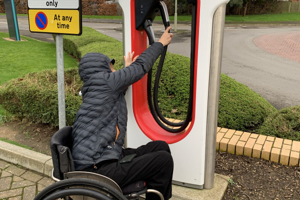 A photo of our Senior Access consultant Tash Davies reaching for a Tesla charger positioned above shoulder height, at the limit of her range of motion, making it difficult to access.