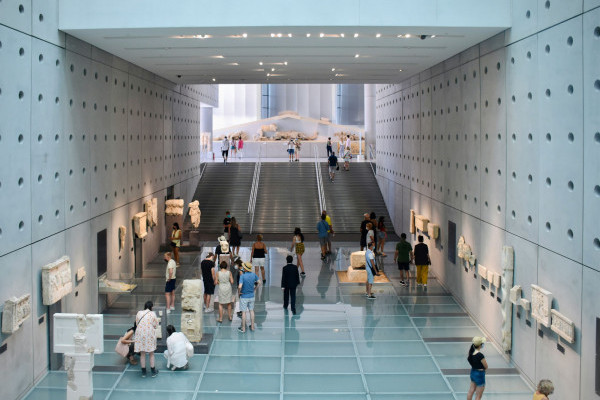 Visitors stand in groups looking at artefacts in a vast open museum. The floor is glass panelled, with concrete walls, displaying carved sculptures. In the view beyond is a wide staircase leading up to another level of the museum.