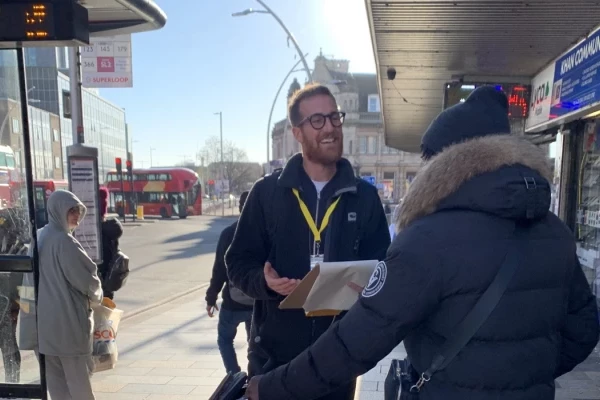 Adam, a member of the team at Mima holds a clipboard whilst interviewing a passenger at a London bus stop in a built up area.