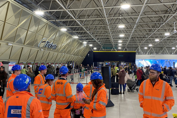 The Mima team on site at Gatwick airport train station wearing high vis orange jackets and blue hard hats. Passengers are moving around the ticket hall with suitcases.