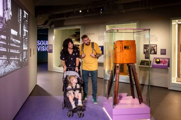 A young family explores a museum gallery featuring historic film and photography equipment. A woman pushes a child in a buggy while a man stands beside them, both looking at a large wooden early film camera displayed in a glass case. Behind them, a wall projection shows a black-and-white film image, and gallery text panels are visible around the room.