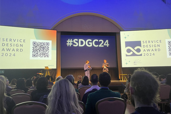 A photo of the main stage at Service Design Global Conference 2024 with two speakers on the stage holding microphones set against the backdrop of a videowall featuring three large screens